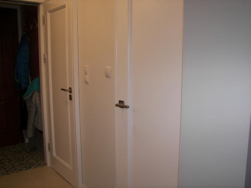 Białe drzwi wewnętrzne z klamkami i włącznikami światła, widoczne ubrania wiszące w otwartym pomieszczeniu po lewej stronie.