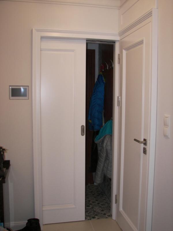 Białe drzwi przesuwne w systemie chowanym, widoczne wnęki na ubrania i wieszaki, minimalistyczny design.