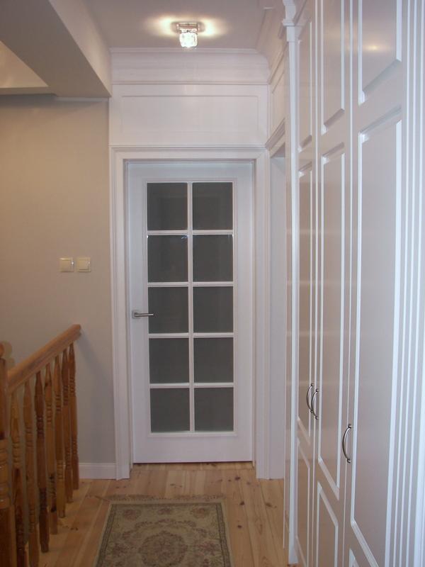 Korytarz z białymi drzwiami z szybkami i zabudowaną szafą wnękową, drewniana podłoga i dywanik, widoczna barierka schodów.