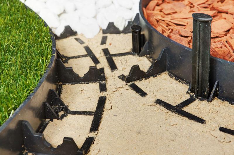 Montaż obrzeża trawnikowego z tworzywa sztucznego, wypełnionego piaskiem, z widocznymi elementami mocującymi i różnymi rodzajami wypełnienia rabat: białym grysem, korą drzewną i trawą.