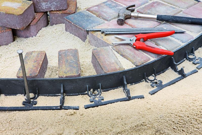 Układanie obrzeży trawnikowych z kostki brukowej: czarny plastikowy obrzeże, młotek, gwoździe i czerwone nożyce na podsypce z piasku.
