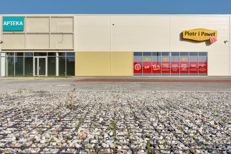 Krata trawnikowa wypełniona drobnym żwirem na parkingu przed budynkiem z szyldami Apteka i Piotr i Paweł.