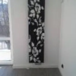 Czarno-biały, dekoracyjny grzejnik panelowy z motywem kwiatowym na białej ścianie w jasnym pomieszczeniu. Widoczny fragment okna i szara podłoga.