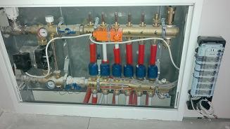 Rozdzielacz ogrzewania podłogowego w białej szafce, z widocznymi rurami w kolorach czerwonym i niebieskim oraz elementami sterującymi, ujęcie z góry.