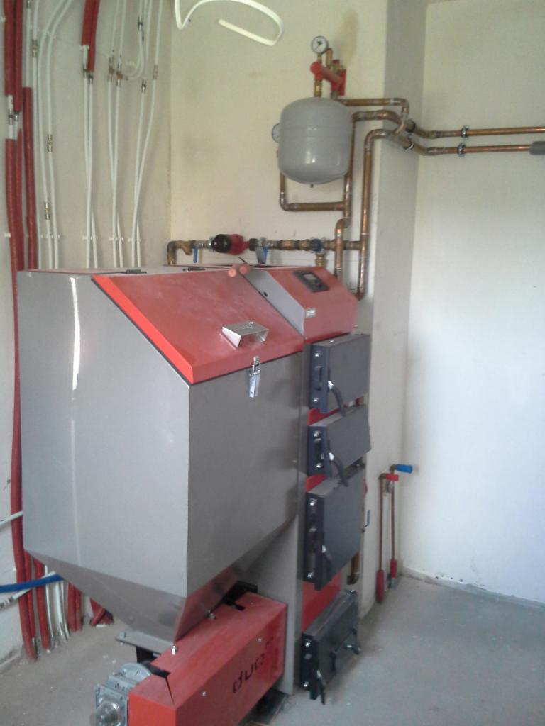 Szary piec CO z czerwonymi akcentami i widoczną instalacją hydrauliczną. Rury miedziane i białe, pionowe przewody. Widok na zainstalowany system grzewczy.