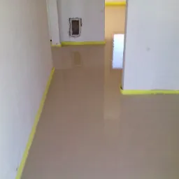 Wylana, gładka posadzka samopoziomująca w pomieszczeniu z białymi ścianami. Żółta taśma zabezpieczająca przy krawędziach. Widoczny otwór w ścianie.