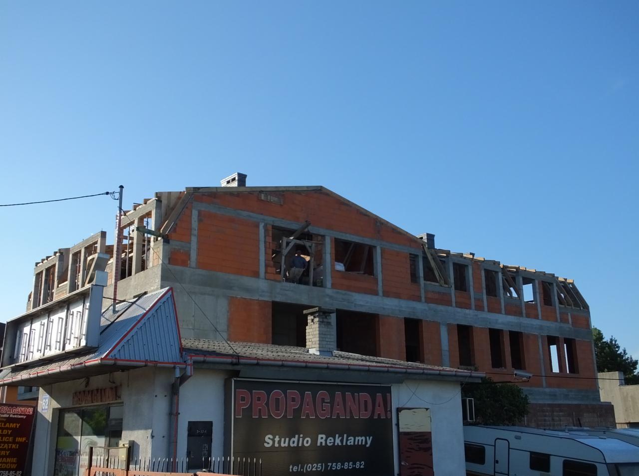 Budowa nowego dachu z czerwonej cegły, widok z dołu na nieukończoną konstrukcję, błękitne niebo w tle. Na pierwszym planie budynek 'Studio Reklamy'.