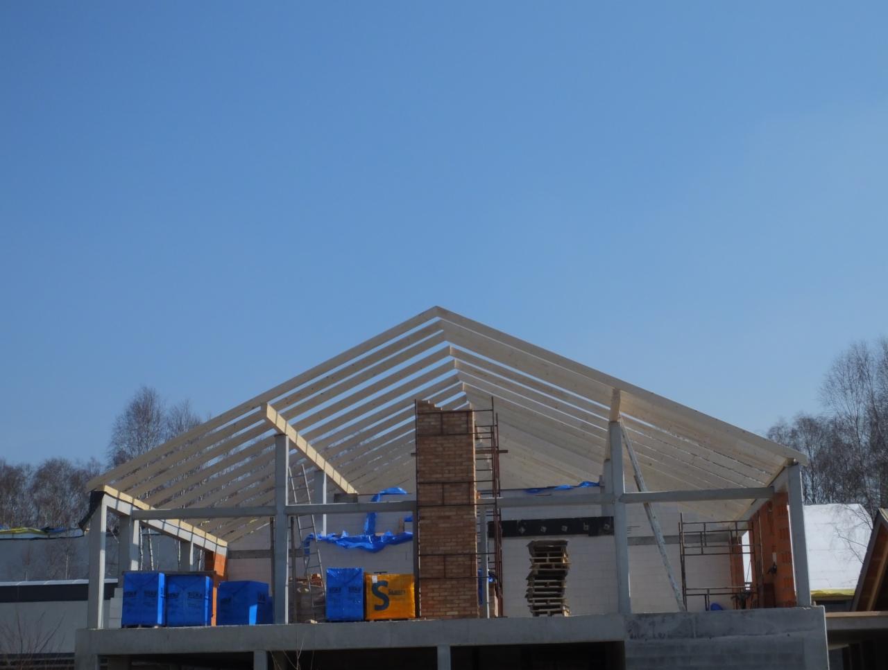 Konstrukcja dachu z drewnianych belek na murowanym budynku w trakcie budowy, widok z dołu na tle błękitnego nieba i drzew.