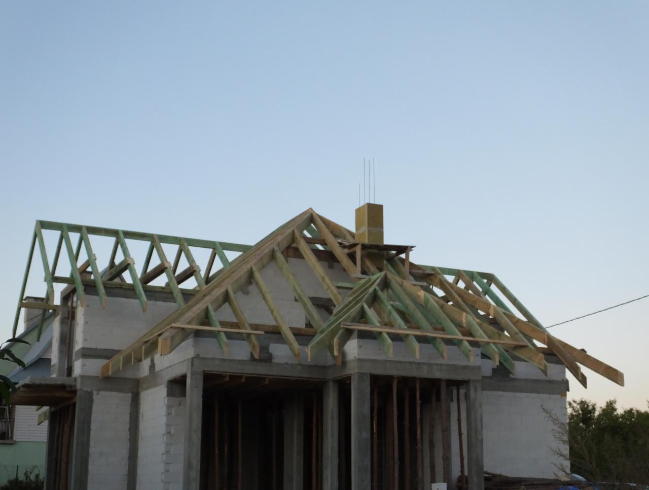 Konstrukcja dachu z drewnianymi belkami na surowym budynku, widok z dołu. W tle błękitne niebo. Etap budowy z kominem w izolacji.