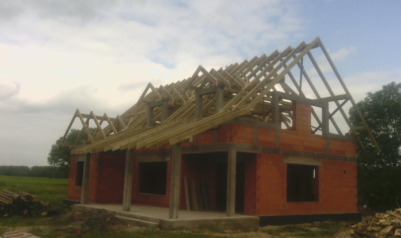 Surowy stan domu z czerwonej cegły z drewnianą konstrukcją dachu na tle pochmurnego nieba i zieleni. Widoczne deski i elementy więźby dachowej.