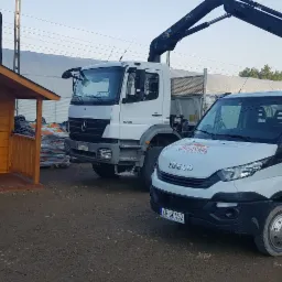 Dostawa ekogroszku: biała ciężarówka Mercedes 1829 z dźwigiem HIAB oraz biała Iveco Soldach-Pol z załadunkiem worków ekogroszku na pace, na tle składu opału z drewnianym domkiem