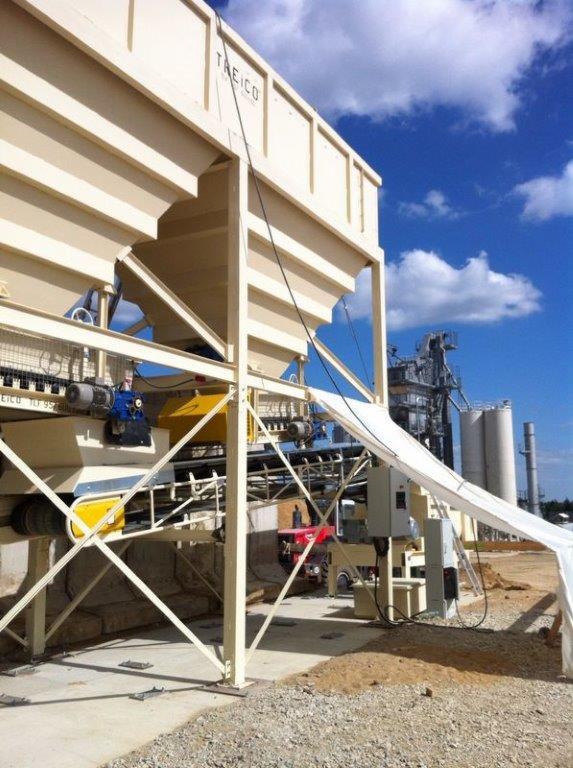 Mieszarka do kruszyw marki TAEICO z systemem przenośników taśmowych i silnikami elektrycznymi, na tle błękitnego nieba z chmurami i widocznej w oddali instalacji przemysłowej z silosami.