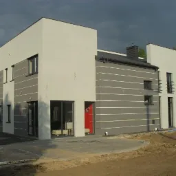 Nowoczesny dom z szarą elewacją i białymi poziomymi pasami, czerwone drzwi wejściowe. Widoczne fragmenty kostki brukowej i teren zielony. Pochmurne niebo w tle.