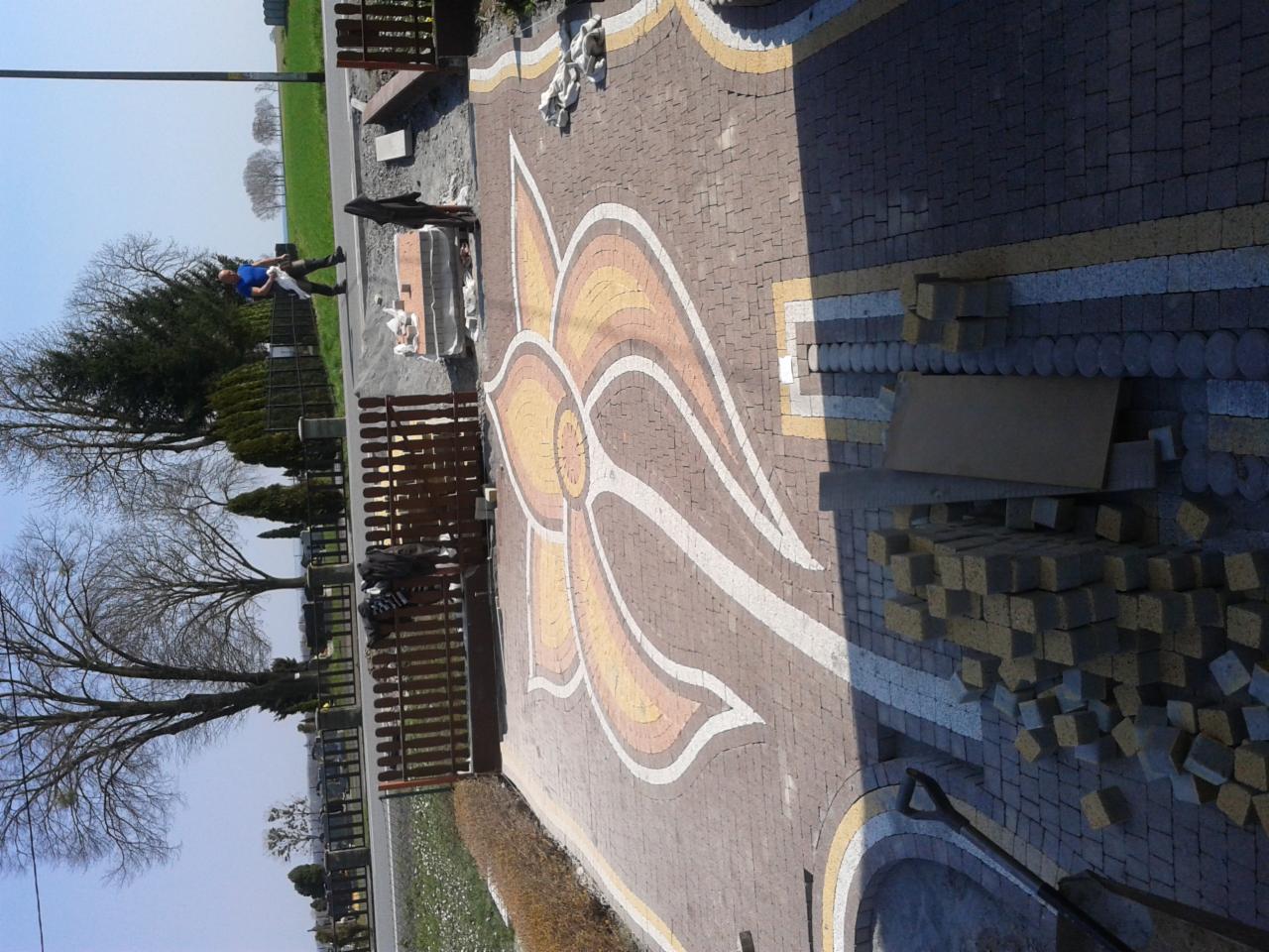 Wzór kwiatowy z kostki brukowej w odcieniach brązu, żółci i bieli. Część kostki jeszcze nieułożona, widoczna łopata i osoby w tle. Ujęcie z góry.