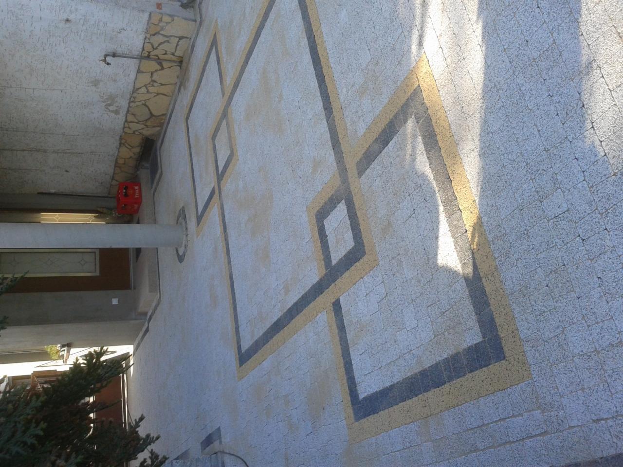 Wzór z kostki brukowej w odcieniach szarości, czerni i beżu, ułożony na podjeździe. Geometryczny motyw z kwadratów. Częściowo zacieniona powierzchnia. Widok z góry.