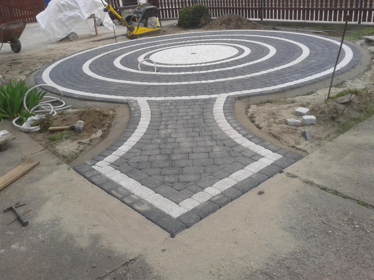 Okrągły wzór z szarej i białej kostki brukowej, tworzący dekoracyjny wzór na podwórku. W tle widać zagęszczarkę i częściowo ułożoną kostkę.
