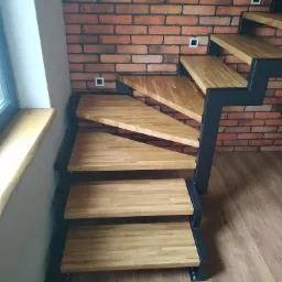 Nowoczesne schody drewniane z metalową konstrukcją, widoczne na tle ściany z cegły i drewnianej podłogi.