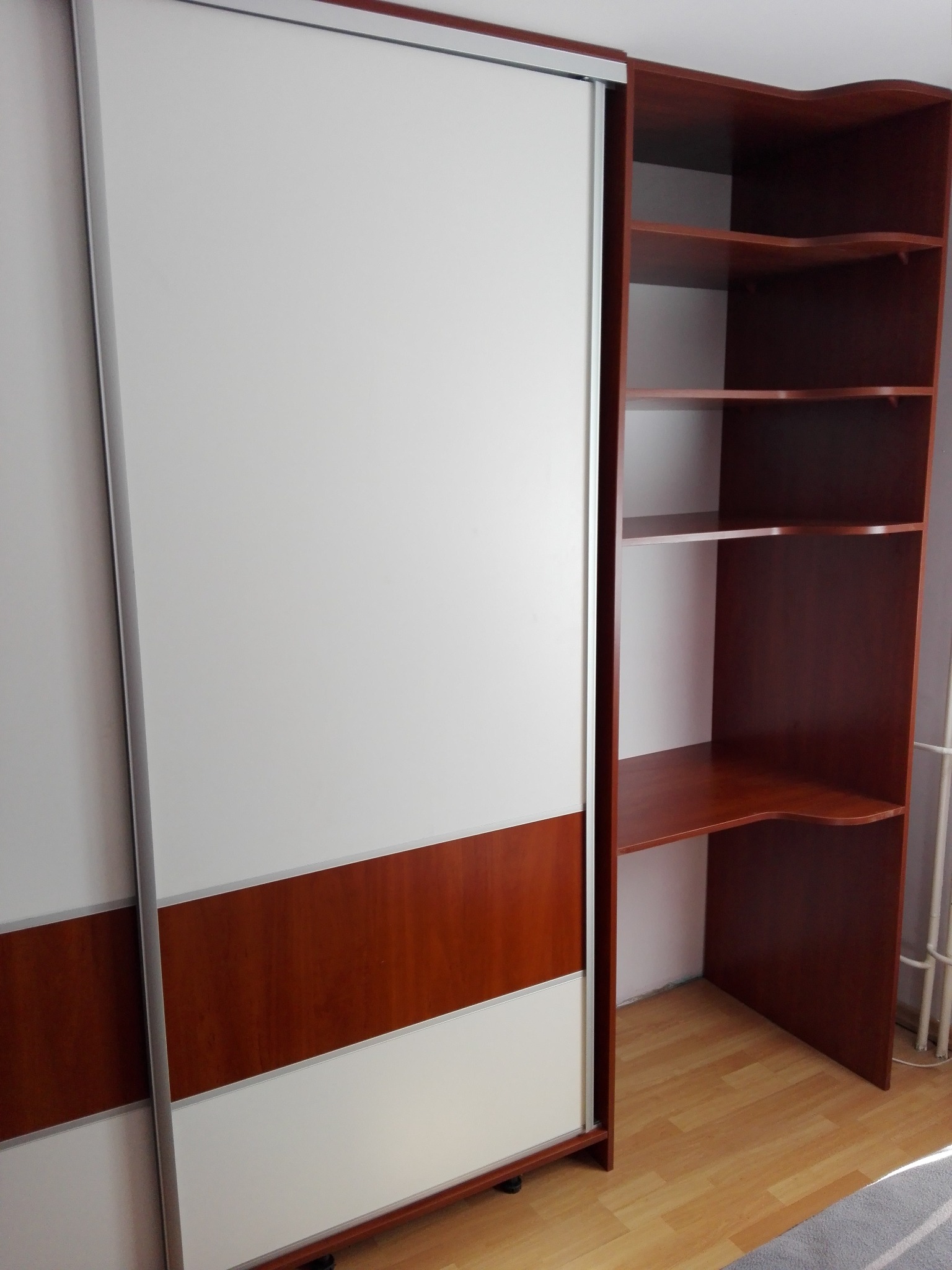 Biało-brązowa szafa przesuwna z półkami w kolorze drewna, z blatem tworzącym narożne biurko, na drewnianej podłodze.