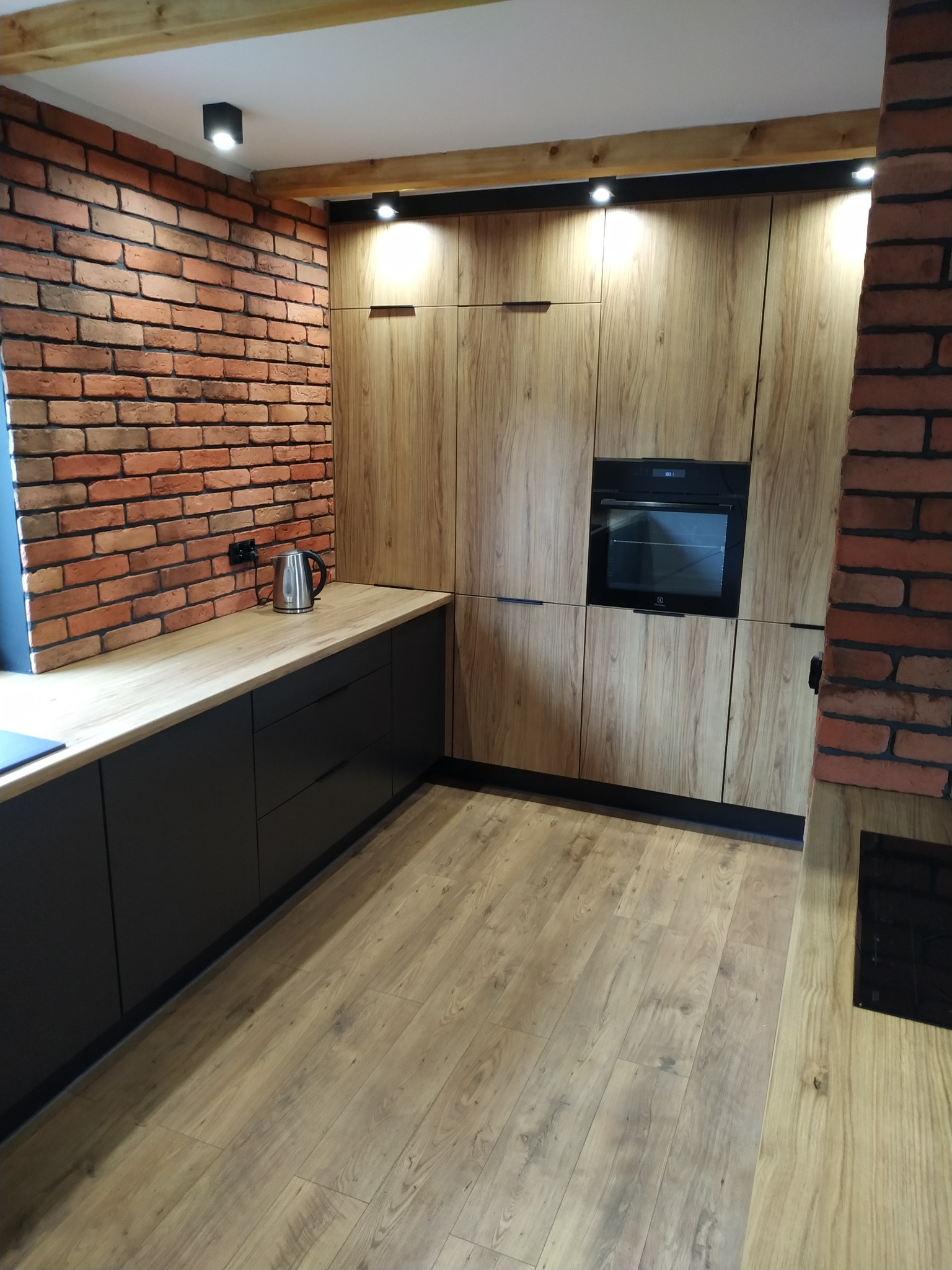 Nowoczesna kuchnia z drewnianymi frontami i czarnymi szafkami, ceglana ściana, drewniany blat, metalowy czajnik i drewniane belki stropowe.