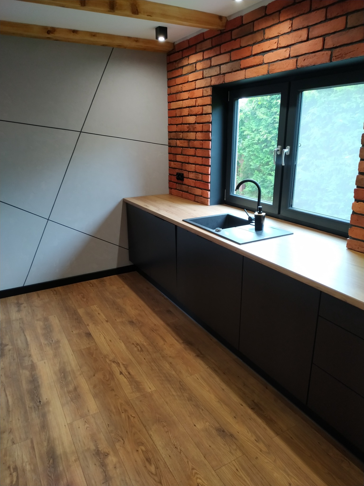 Nowoczesna kuchnia z czarnymi szafkami, drewnianym blatem i ceglaną ścianą, widok na zewnątrz przez okno.