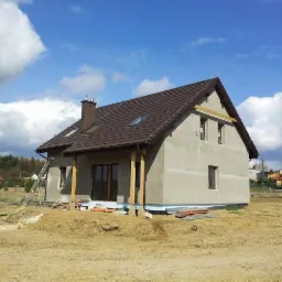 Surowy stan elewacji domu jednorodzinnego z brązowym dachem, widoczna drabina oparta o ścianę, przygotowany teren wokół budynku.