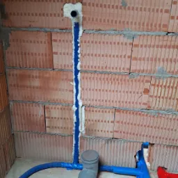 Instalacja wodno-kanalizacyjna w surowym stanie: niebieskie rury przytwierdzone do ceglanej ściany, szara rura odpływowa, pianka montażowa uszczelniająca przejścia.