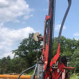 Drill-Team Bartłomiej Kuźmicki - Czerwona wiertnica ROTOMAX XL GTC na tle błękitnego nieba z chmurami i zieleni drzew, z operatorem w tle.