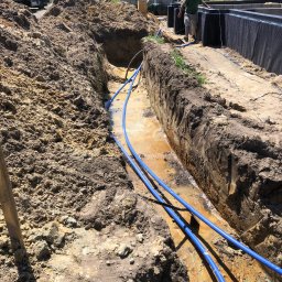 Drill-Team Bartłomiej Kuźmicki - Wykop z ułożonymi niebieskimi rurami, prawdopodobnie instalacja podziemna, widoczny fragment budynku i pracownik w tle.