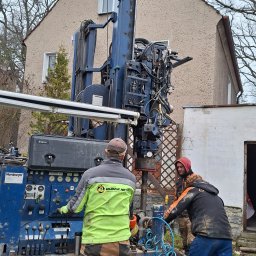 Drill-Team Bartłomiej Kuźmicki - Wiertnica Nordmeyer w trakcie pracy przy budynku jednorodzinnym, dwóch operatorów w roboczych ubraniach obsługuje maszynę, widoczna instalacja hydrauliczna i manometry.