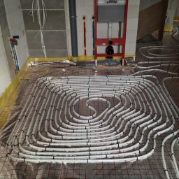 Drill-Team Bartłomiej Kuźmicki - Rozłożone rury ogrzewania podłogowego na folii izolacyjnej z siatką, widoczne fragmenty ścian w surowym stanie i instalacje sanitarne.
