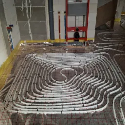 Rozłożone rury ogrzewania podłogowego na folii izolacyjnej z siatką, widoczne fragmenty ścian w surowym stanie i instalacje sanitarne.