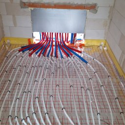 Drill-Team Bartłomiej Kuźmicki - Rozłożona instalacja ogrzewania podłogowego w surowym pomieszczeniu z białych bloczków, widoczne rury w kolorze białym, czerwonym i niebieskim, przymocowane do siatki z czerwonymi kwadratami.