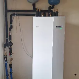 Biała jednostka wewnętrzna z szarym panelem bocznym, zamontowana w pomieszczeniu z widocznymi przewodami, zaworami i trzema zbiornikami wyrównawczymi na ścianie.