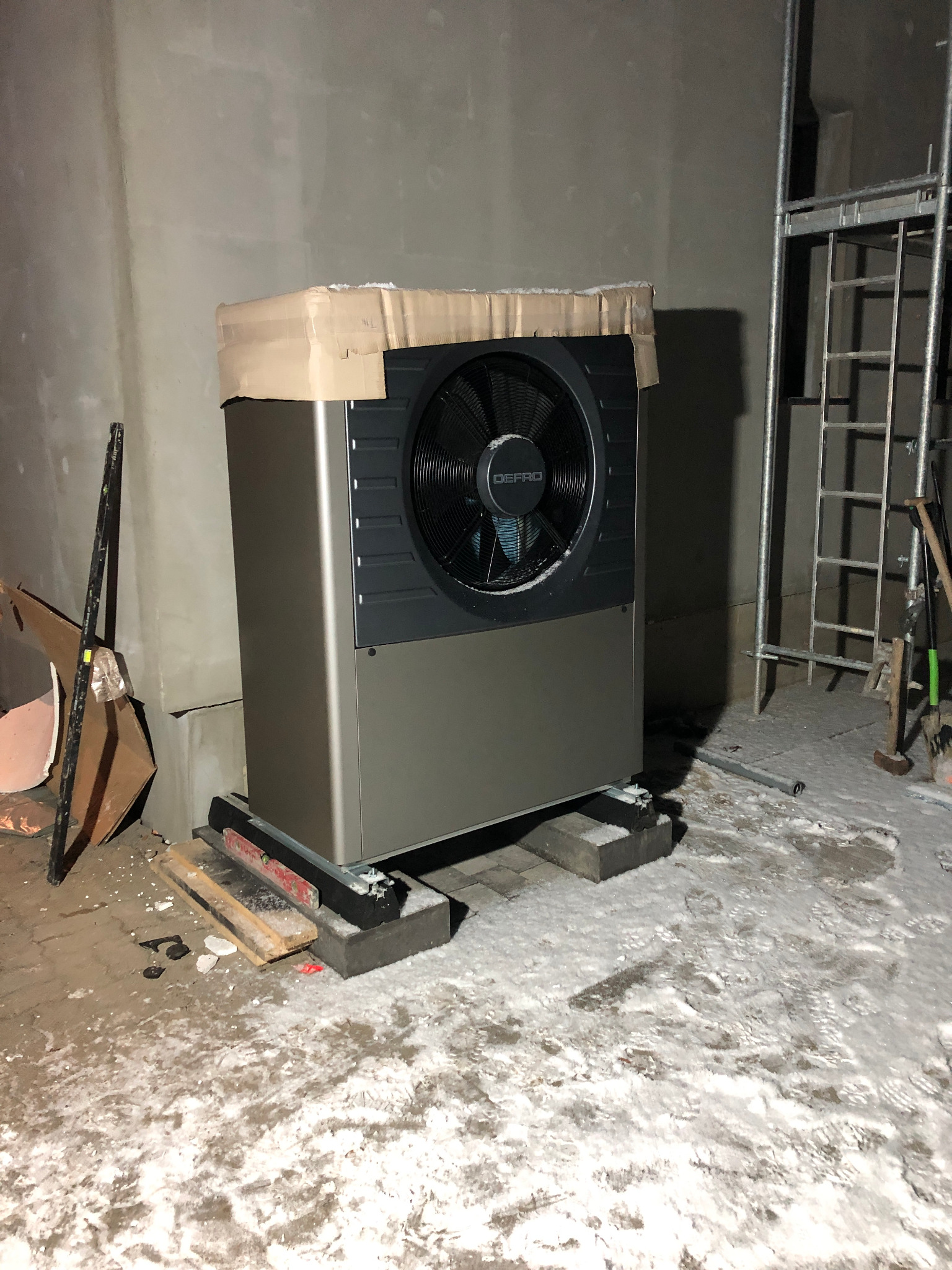 Szaro-czarna jednostka zewnętrzna urządzenia grzewczego marki Defro, tymczasowo zabezpieczona kartonem, umieszczona na betonowych bloczkach w surowym, ośnieżonym pomieszczeniu w trakcie montażu,...