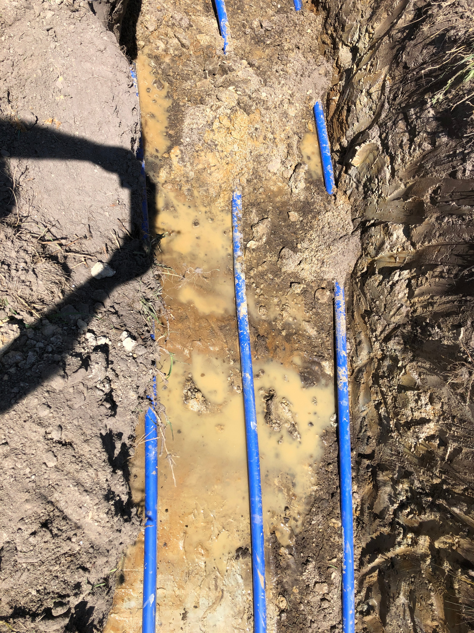 Wykop z niebieskimi rurami w Kamieńcu Wrocławskim, częściowo zalany wodą, cień osoby po lewej stronie.