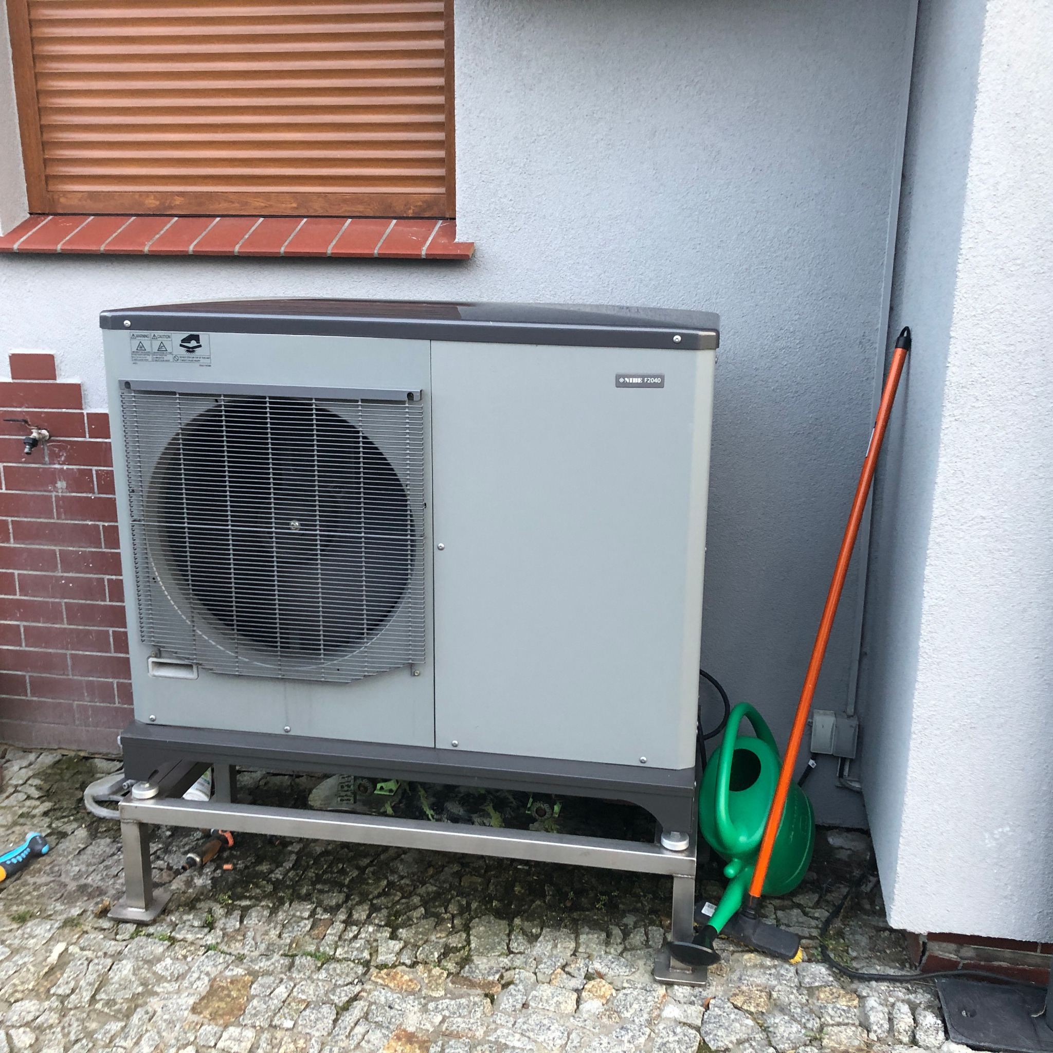 Szara pompa ciepła NIBE F2040 na metalowej podstawie obok ściany domu z oknem, obok stoi pomarańczowy kij i zielona konewka.