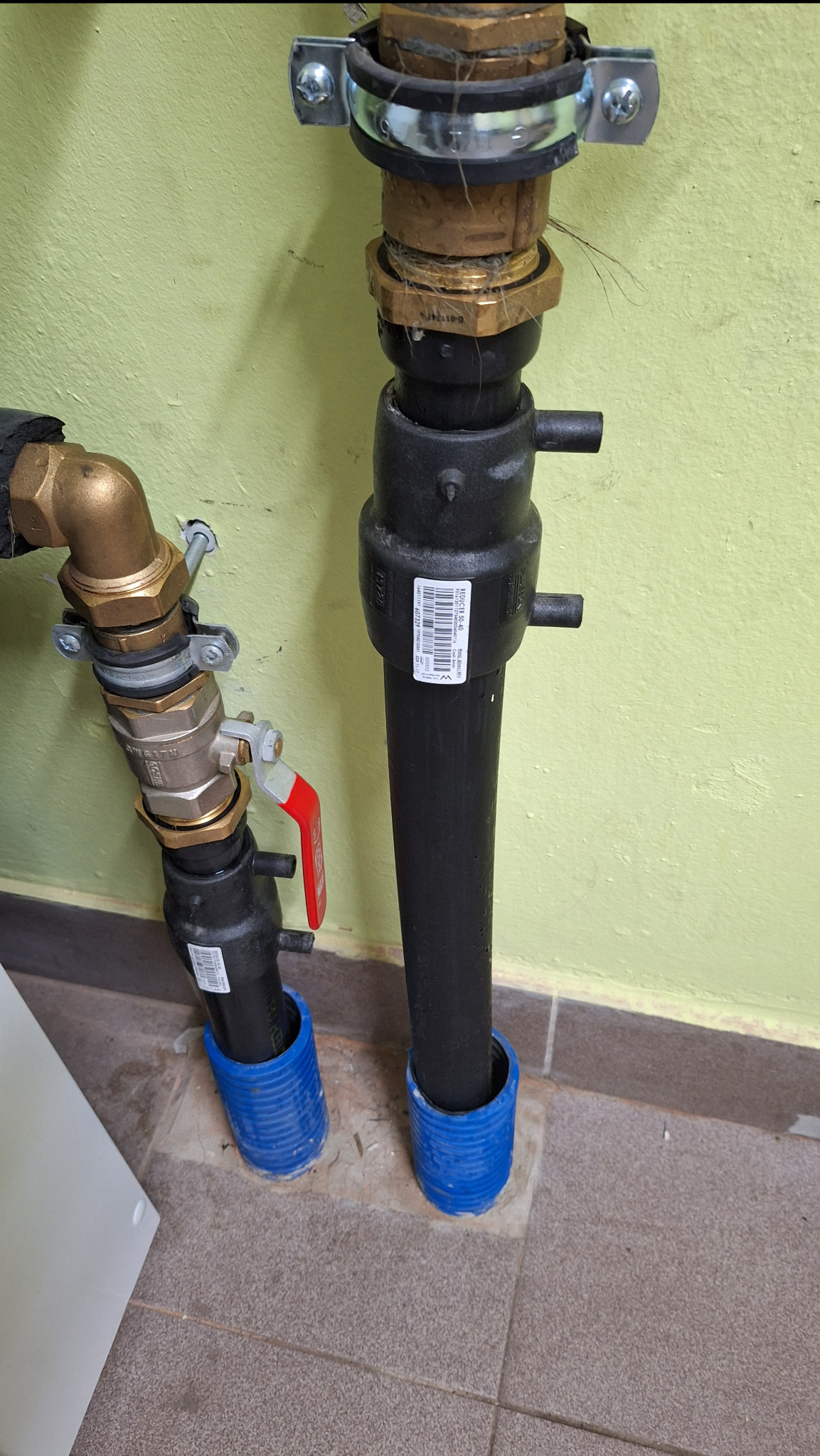 Dwie rury instalacyjne, jedna z zaworem z czerwoną rączką, wystające z podłogi zabezpieczone niebieskimi osłonami, widoczne złączki i mocowania do ściany w jasnym odcieniu zieleni.