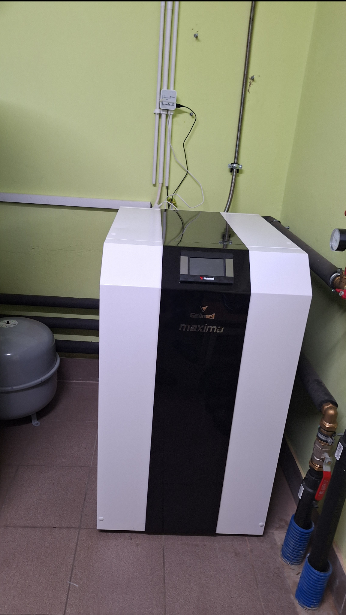 Biało-czarna obudowa urządzenia Galmet Maxima z wyświetlaczem, rury instalacyjne w tle, szara ściana i brązowe płytki podłogowe.