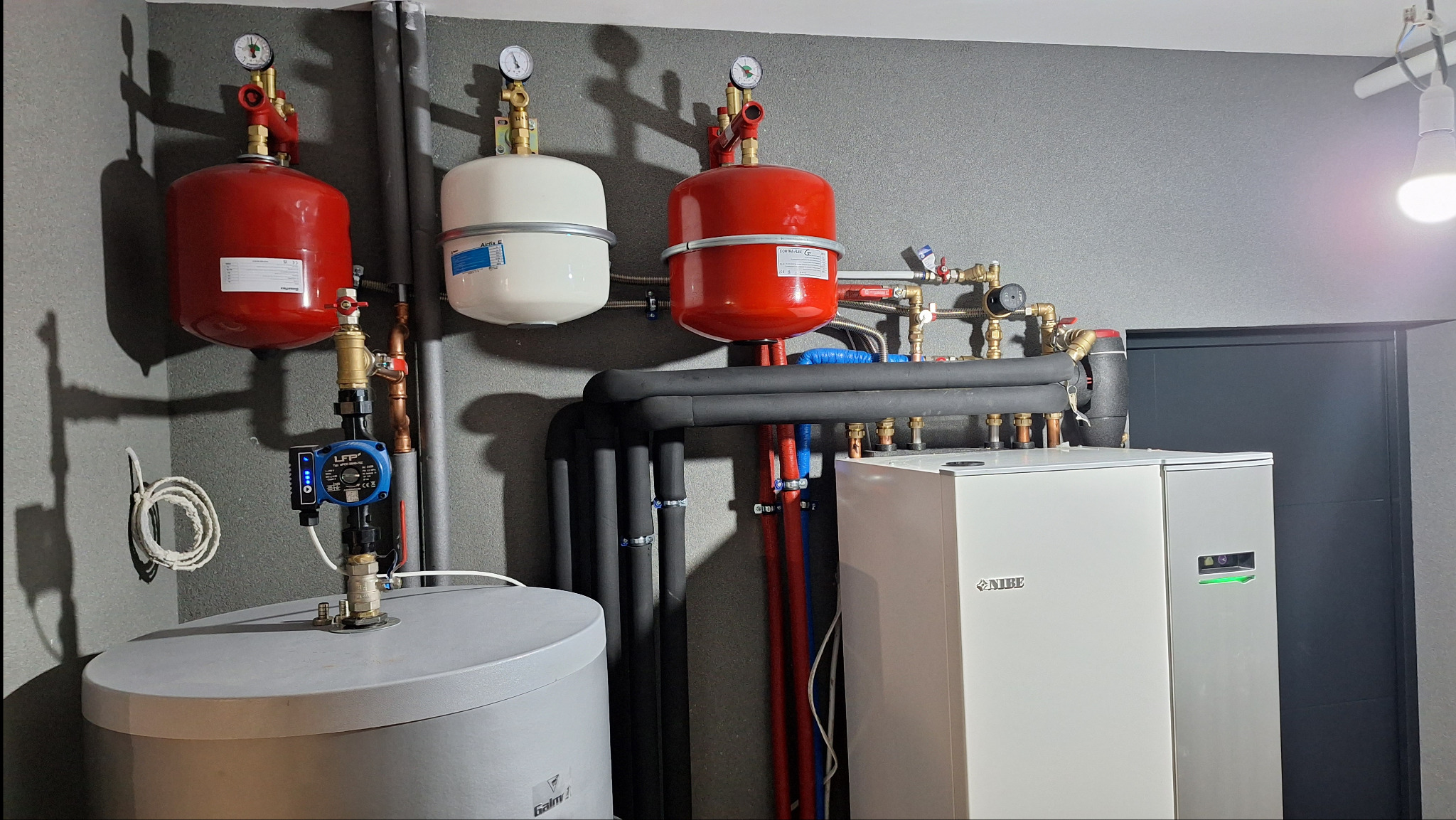 Instalacja grzewcza w kotłowni: dwa czerwone i jeden biały zbiornik wyrównawczy, pompa obiegowa LFP, grube czarne rury izolacyjne i nowoczesny piec grzewczy NIBE z zielonym podświetleniem, szara...