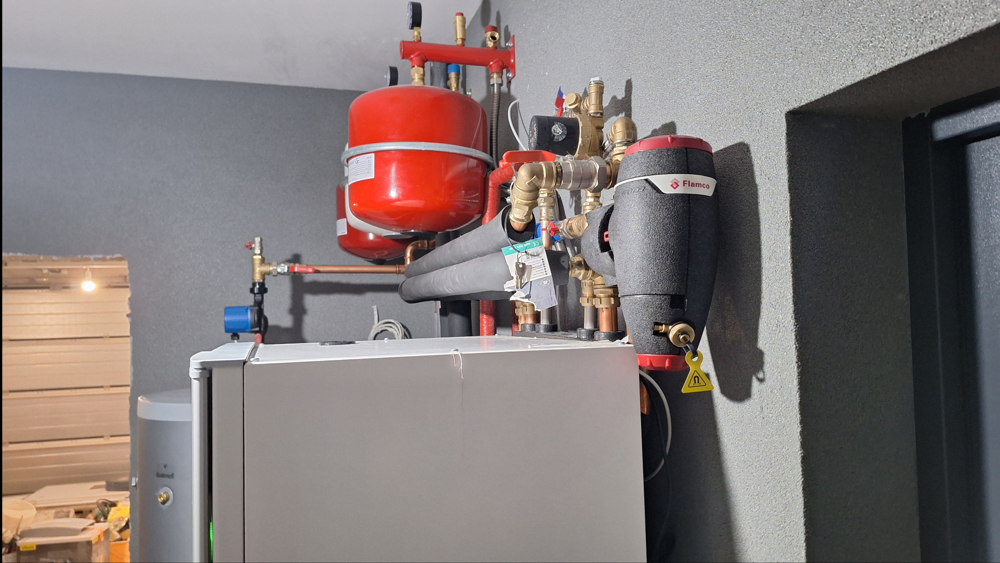 Zainstalowany system grzewczy: czerwony zbiornik wyrównawczy, czarny separator powietrza Flamco, rury izolowane termicznie, zawory i biała obudowa kotła na tle szarej ściany.