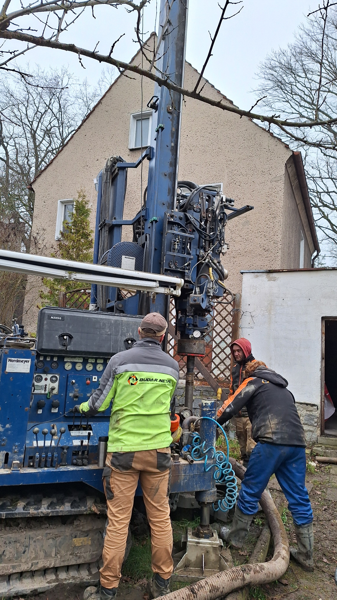 Wiertnica Nordmeyer w trakcie pracy przy budynku jednorodzinnym, dwóch operatorów w roboczych ubraniach obsługuje maszynę, widoczna instalacja hydrauliczna i manometry.
