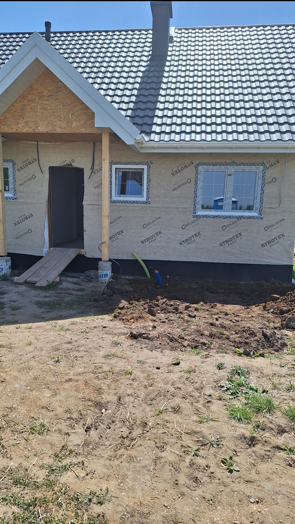 Dom w trakcie budowy z ociepleniem ścian zewnętrznych, widoczne rury wystające z ziemi, przygotowane pod instalację zewnętrzną.