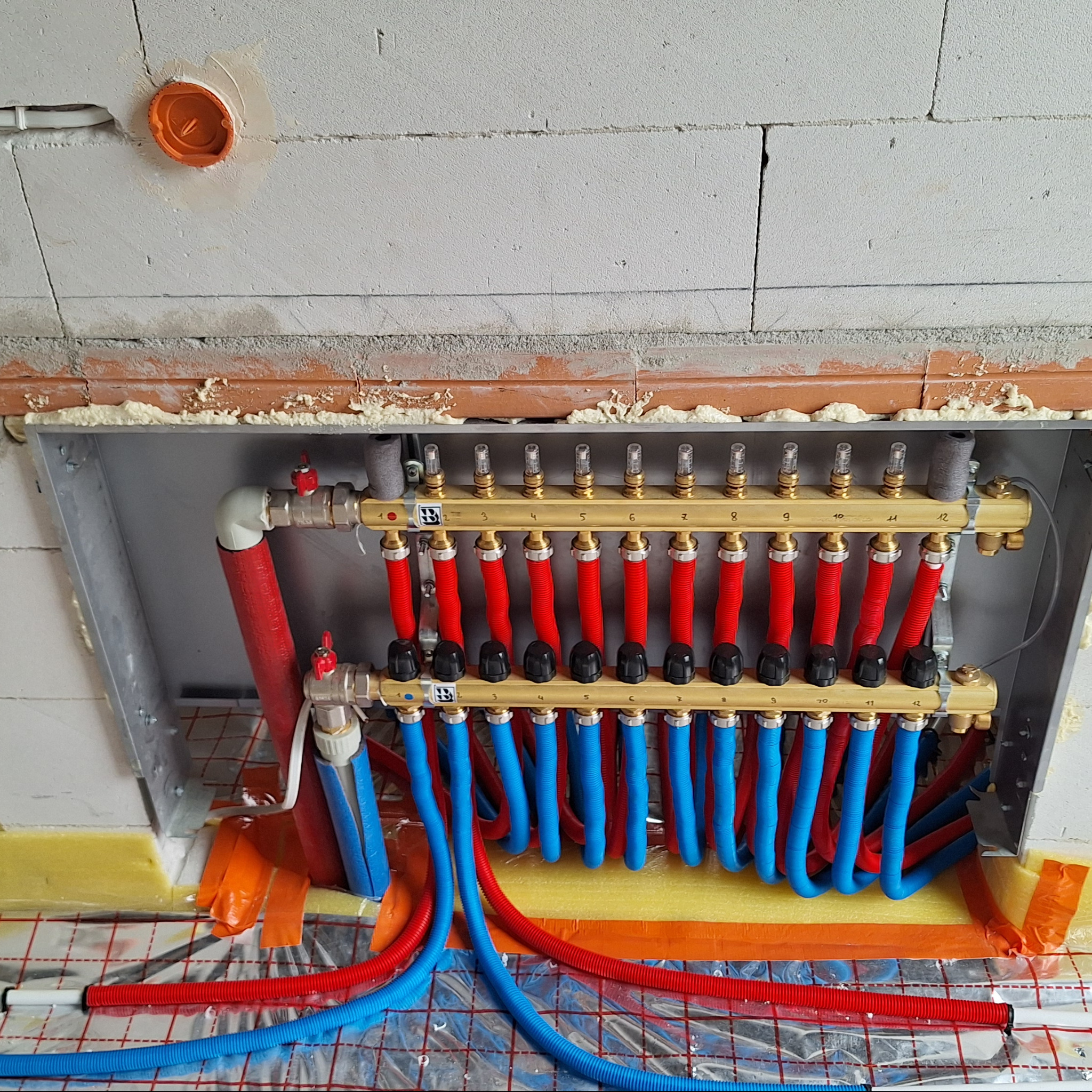 Rozdzielacz ogrzewania podłogowego z rurami w kolorze czerwonym i niebieskim, zamontowany w ścianie z pustaków, widoczne izolacje i folia grzewcza.
