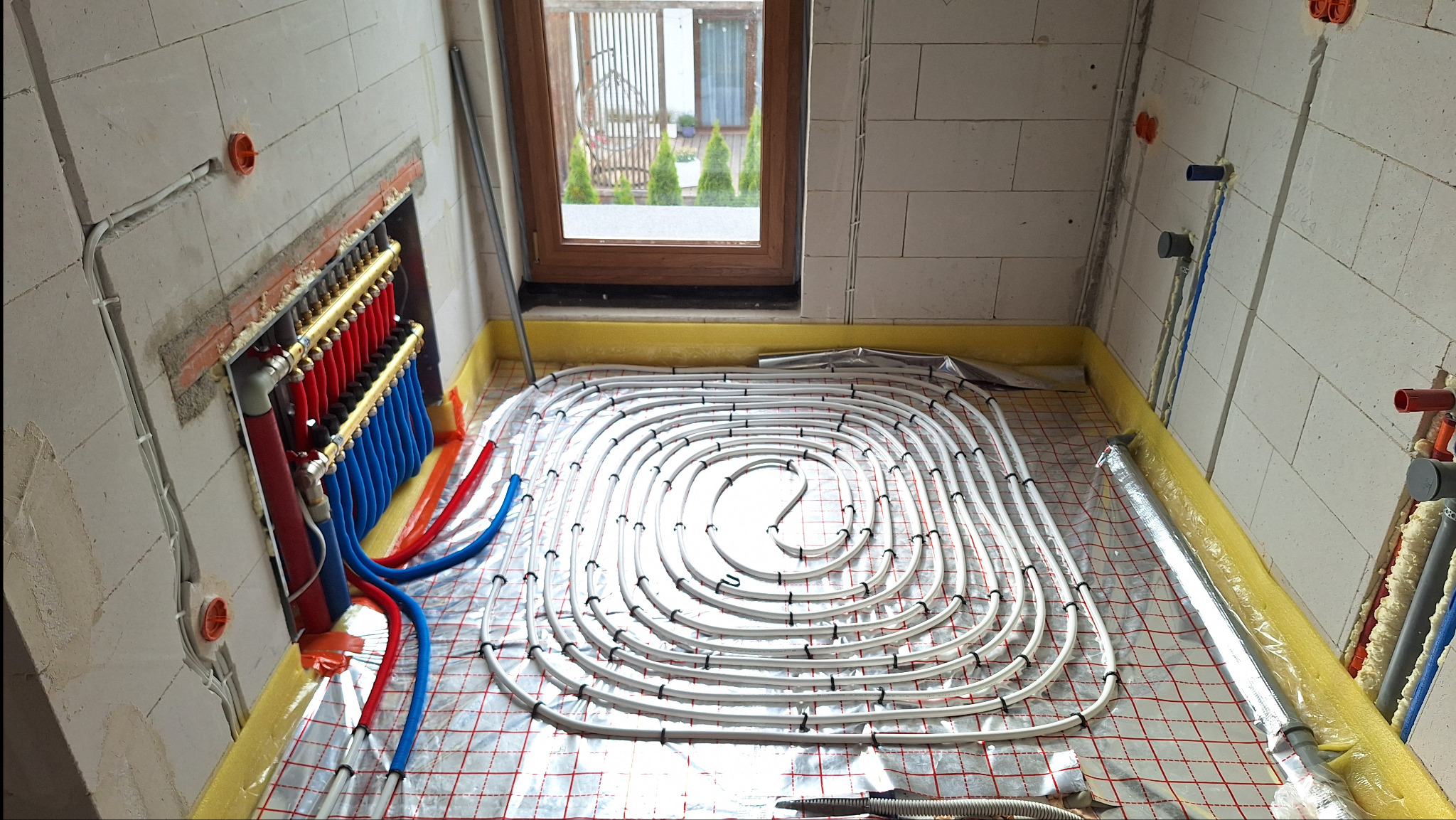 Rozłożone rury ogrzewania podłogowego na folii z kratką, widoczny rozdzielacz z czerwonymi i niebieskimi rurami, okno z widokiem na zewnątrz i surowe ściany z instalacją elektryczną.