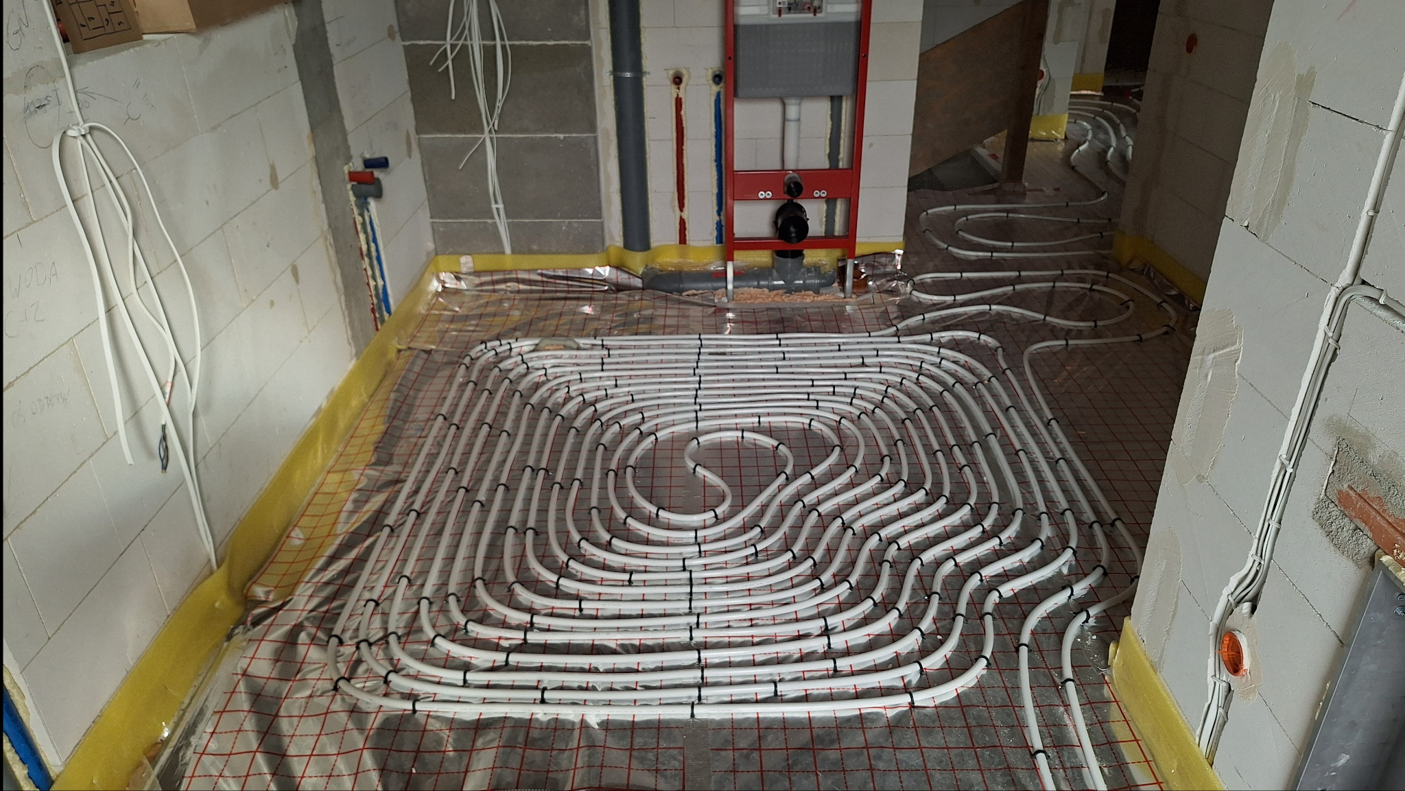 Rozłożone rury ogrzewania podłogowego na folii izolacyjnej z siatką, widoczne fragmenty ścian w surowym stanie i instalacje sanitarne.