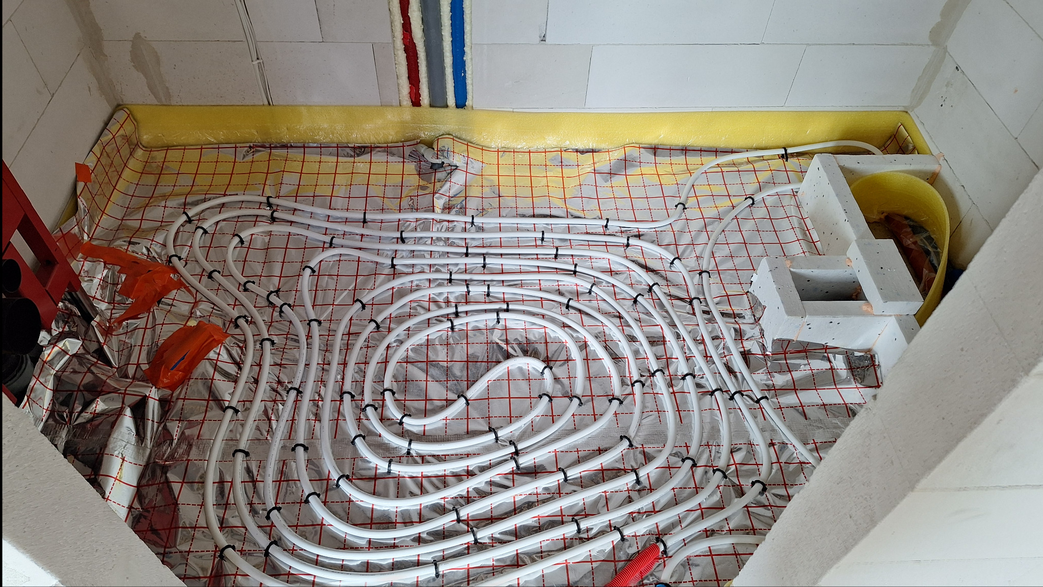 Instalacja ogrzewania podłogowego z białych rur na folii z czerwoną kratką, widok z góry na surowy stan budynku, fragment drabiny malarskiej w lewym rogu.