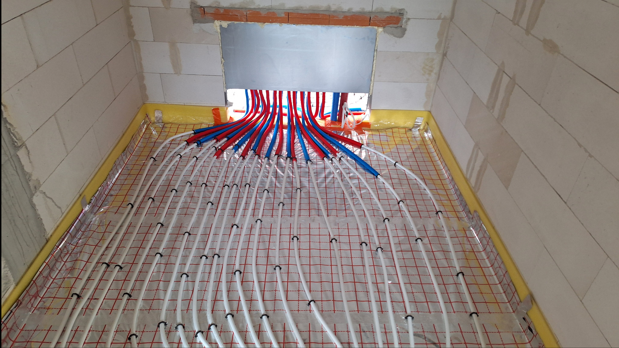 Rozłożona instalacja ogrzewania podłogowego w surowym pomieszczeniu z białych bloczków, widoczne rury w kolorze białym, czerwonym i niebieskim, przymocowane do siatki z czerwonymi kwadratami.