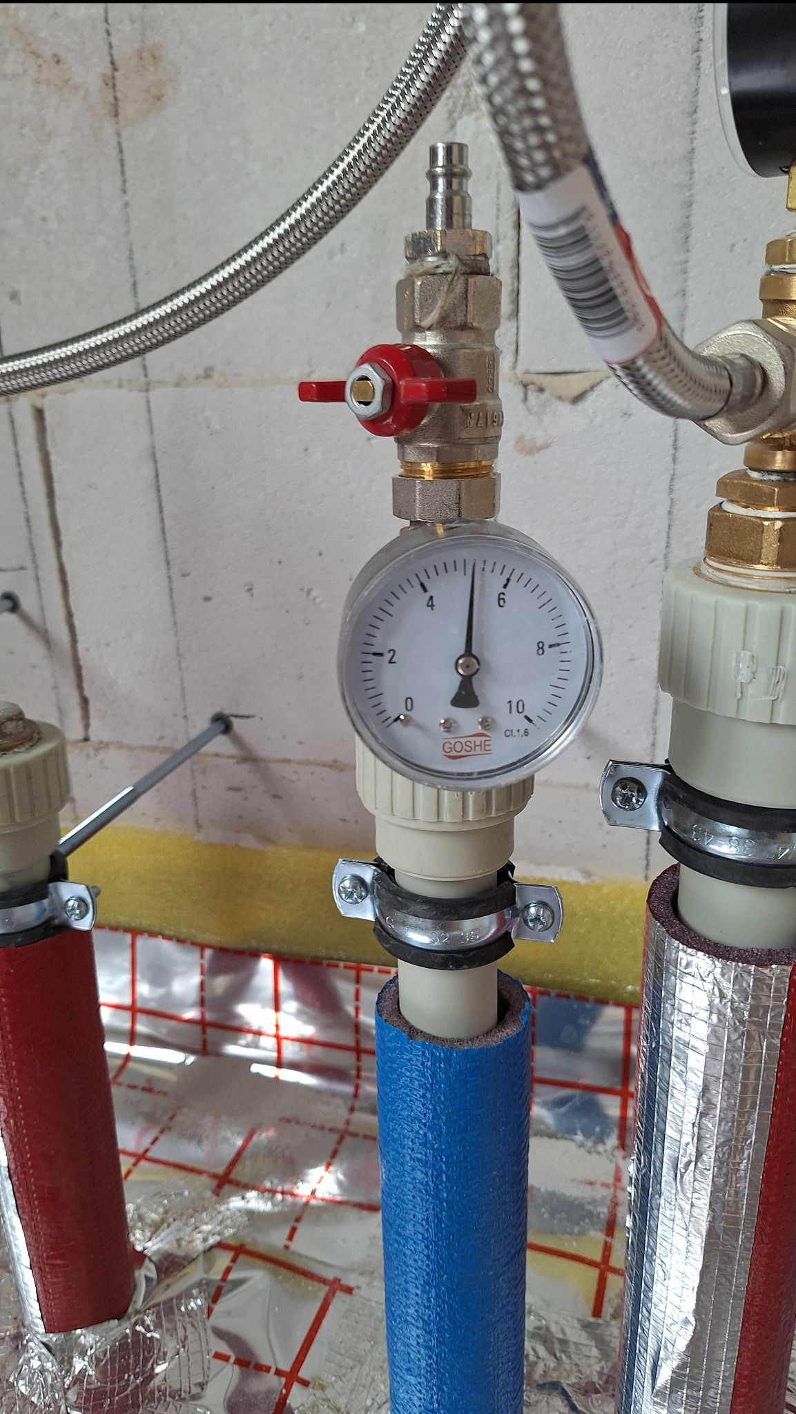 Instalacja grzewcza z manometrem Goshe wskazującym ciśnienie, rury w izolacji czerwonej, niebieskiej i srebrnej, zawór z czerwoną rączką, podłogówka w tle.