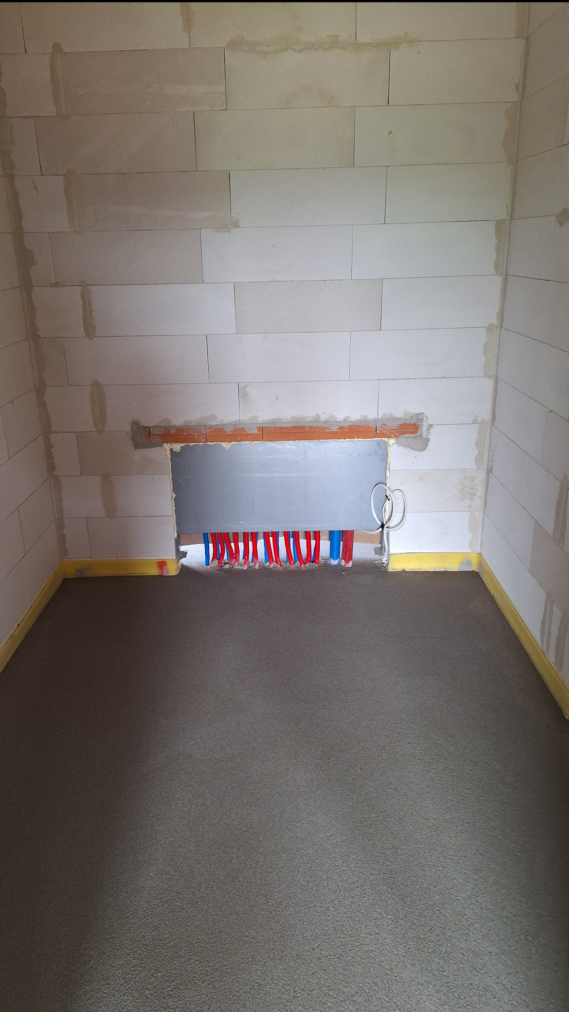 Wnętrze w trakcie budowy z widocznymi rurami ogrzewania podłogowego wystającymi ze ściany z betonu komórkowego, świeżo wylana posadzka.