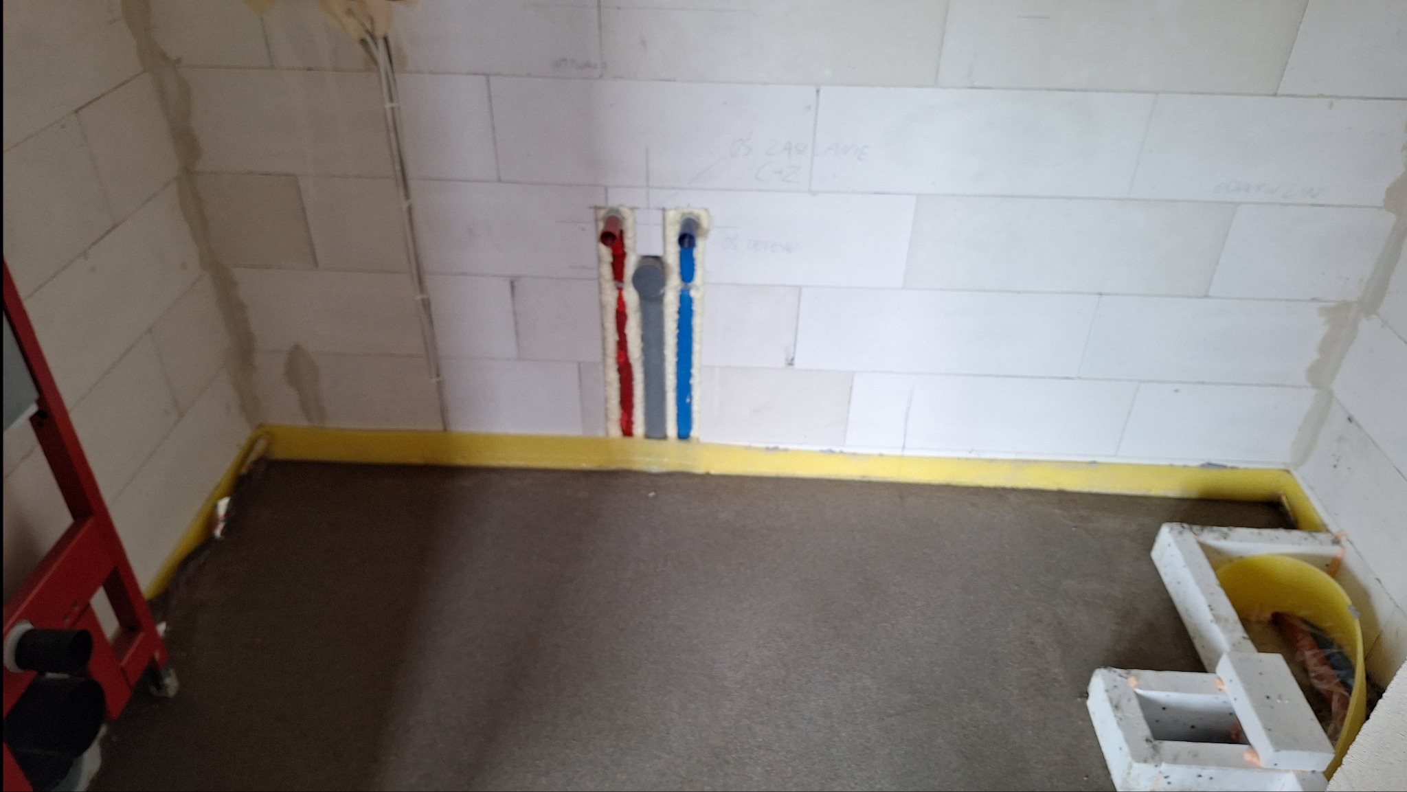 Przygotowanie podłoża pod instalację ogrzewania podłogowego: wylewka cementowa, zabezpieczone ściany taśmą, widoczne wyprowadzenia rur instalacyjnych w ścianie z betonu komórkowego.